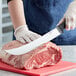 A person in gloves using a Schraf cimeter knife to cut raw meat on a cutting board.