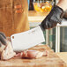 A hand in a black glove uses a Schraf cleaver to cut a chicken leg on a cutting board.