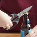 A hand uses Mercer Culinary German steel kitchen shears to cut a bottle of beer.