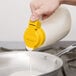 A close-up of a Tablecraft 48 oz. dispenser jar with a yellow top being used to pour a liquid into a pan.