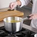 A person in a white coat using a Choice aluminum sauce pan to pour soup into on a stove.