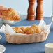 A hand holding a croissant in a Tablecraft natural-colored oblong bread basket.