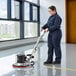 A woman in a blue jumpsuit using a Lavex single speed floor machine to clean a floor.