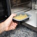 A hand holding a Pactiv Newspring black rectangular VERSAtainer filled with food in front of a microwave.