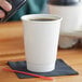 A hand holding a white Choice double wall paper hot cup with a lid over coffee on a napkin.