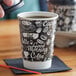 A hand pours coffee into a black and white Choice Coffee Break paper cup on a table.