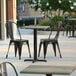 A Lancaster Table & Seating square table top with a textured metal finish on a table with black chairs outside.