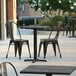 A black Lancaster Table & Seating square table top with a smooth finish on an outdoor patio with black metal chairs.