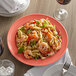 A Tuxton Concentrix Cinnebar china plate with shrimp pasta and basil on a table with a fork and a glass of wine.