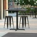 A Lancaster Table & Seating counter height table with a grey surface and cross base with three black stools on a patio.