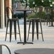 A Lancaster Table & Seating bar height table with a smooth grey surface and metal base plate on a sidewalk with three black bar stools.