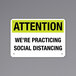 A black and yellow rectangular decal on a wall that says "Attention / We're Practicing Social Distancing" with black text.