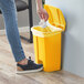 A person's leg putting a white bag in a yellow Lavex rectangular step-on trash can.