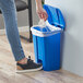 A hand putting a white bag into a blue Lavex step-on trash can.