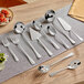 Acopa Edgeworth 10-piece stainless steel serving utensils set on a table with silverware including a spoon and knife.