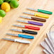A group of Choice serrated paring knives with colored handles on a wooden surface.