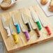 A group of Choice bread knives with yellow, blue, and purple handles on a cutting board.