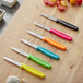 A Choice serrated edge paring knife with a neon yellow handle on a wooden cutting board with other colorful knives.