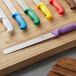 A Choice serrated bread knife with a yellow handle on a wood cutting board with other knives.