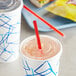 A plastic cup with a red straw in it on a plate with another cup and a sandwich.