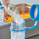 A person wearing a glove pours ice from a metal scoop into a clear container using a blue plastic Vigor Polar Paddle.