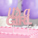 A pink "It's a Girl" cake topper on a pink cake.