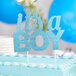 A close up of a white cake with blue frosting and a blue glittered "It's a Boy" cake topper.