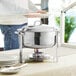 A Choice Deluxe stainless steel soup chafer with silver accents on a table with plates and silverware.