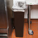 A Continental brown rectangular wall hugger trash can on a metal surface next to a stainless steel sink.