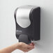 A hand holding a San Jamar black and silver manual foam hand soap and sanitizer dispenser.