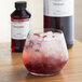 A glass of red liquid with ice and a bottle of LorAnn Pomegranate Super Strength Flavor on a table.