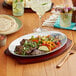 A Choice oval aluminum sizzler platter with food on a table with a drink and a fork.