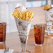 A Carnival King paper cone of french fries on a table with drinks and a newspaper holder.