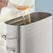 A hand pouring brown liquid into an Avantco stainless steel iced tea dispenser.