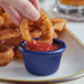 A hand holding a fried onion ring and dipping it into a blue Acopa fluted ramekin of ketchup.