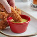 A person dipping a fried chicken strip into a red Acopa ramekin of sauce.