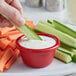 A person dipping a cucumber into a red Acopa ramekin of white sauce.