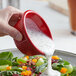 A person pouring white dressing into a bowl of salad using a red fluted ramekin.