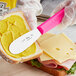 A person using a Choice stainless steel sandwich spreader with a neon pink handle to spread butter on a sandwich.