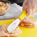 A hand with a yellow gloved holding a Choice stainless steel sandwich spreader over a yellow cutting board with food.