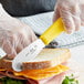 A person in gloves using a Choice stainless steel sandwich spreader to cut a sandwich.