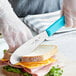 A person using a Choice stainless steel sandwich spreader with a neon blue handle to cut a sandwich.