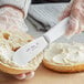 A person using a Choice stainless steel sandwich spreader to spread butter on a bagel.