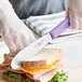 A person using a Choice purple scalloped sandwich spreader to cut a sandwich.