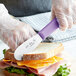 A person using a Choice Scalloped Sandwich Spreader with a purple handle to cut a sandwich.