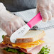 A person using a Choice stainless steel sandwich spreader with a neon pink handle to cut a sandwich.