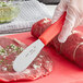 A gloved hand using a Choice stainless steel scalloped sandwich spreader to cut meat.