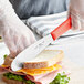 A person using a Choice stainless steel sandwich spreader with a red handle to cut a sandwich.