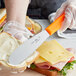 A person's hand using a Choice scalloped sandwich spreader with a neon orange handle to spread butter on a sandwich.