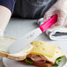 A person holding a Choice stainless steel sandwich spreader with a neon pink handle spreading butter on a sandwich.